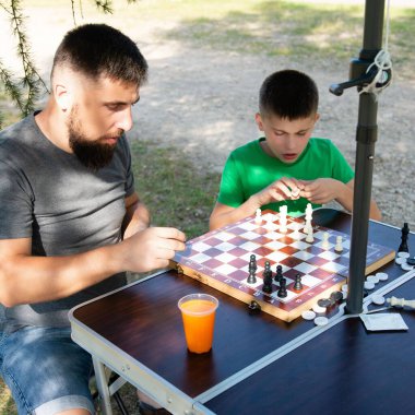 Bir adam ve bir çocuk portatif bir masada açık havada satranç oynuyorlar. İkisi de dikkatle oyuna odaklanmış, park benzeri bir ortamda paylaşılan bir aktivitenin tadını çıkarıyorlar..