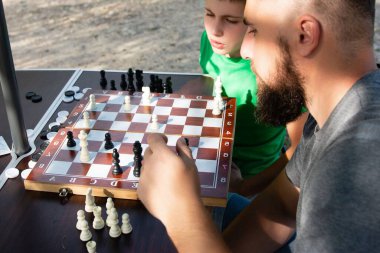 Bir satranç tahtasına odaklanmış, bir yetişkin ve bir çocuk satranç oynuyorlar. Ortam açık, muhtemelen bir park ya da eğlence alanı..