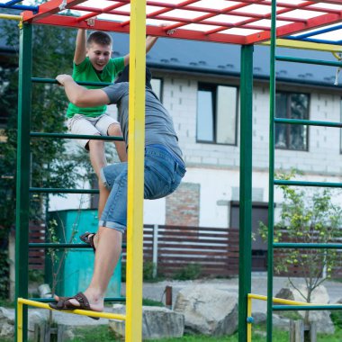 İki genç çocuk renkli bir açık hava tırmanma iskeletine neşeyle tırmanıyor. Bir çocuk diğerine yardım ediyor. Güneşli bir günde bir evin yanında eğleniyorlar..