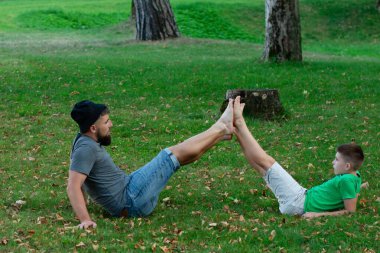 Bir adam ve bir çocuk çimenli bir parkta neşeli bir egzersiz rutinine girişiyor. Birbirlerinin bacaklarını şakayla tutarak eğlenceli bir fitness aktivitesi sergiliyorlar..