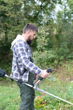 Açık havada, ekose gömlek giyen sakallı bir adam bahçedeki çimleri biçmek için tel makası kullanıyor. Ağaçlar ve yapraklar, güneşli bir günde yemyeşil bir zemin oluşturur..