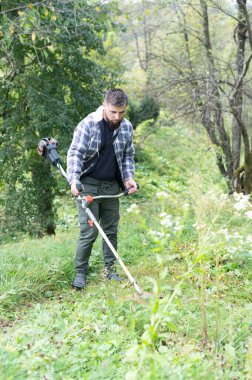 Sakallı bir adam çimenli bir tarlada çim biçme makinesi kullanırken görülüyor. Ağaçlar ve yapraklarla çevrili. O, ekose gömlek, bir tişört ve koyu renk pantolondan oluşan günlük bir kıyafet giydi..