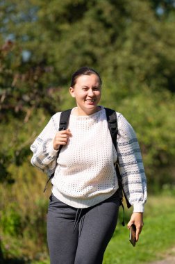 Sırtında desenli bir süveter ve sırt çantası olan gülümseyen bir kadın elinde telefonla güneşli yeşil bir parkta yürüyor ve açık havada aktif bir günün tadını çıkarıyor..