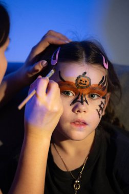 Genç bir kız Cadılar Bayramı tasarımıyla yüzünü boyatıyor. Yetişkin biri, sırtında balkabağı olan bir örümcek yaratmak için turuncu, siyah ve beyaz boya kullanır..