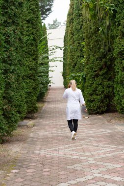 Beyaz önlüklü bir kadın yeşil ağaçlarla çevrili tuğla bir yolda yürüyor. Sahne, bu doğal ortamda sükuneti ve mahremiyeti çağrıştırıyor. Yol bir binaya çıkıyor..