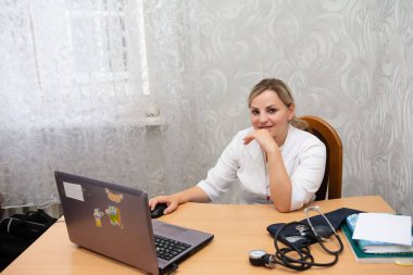 Beyaz laboratuvar önlüklü bir tıp uzmanı bir masada oturuyor, bir dizüstü bilgisayar üzerinde çalışıyor ve bir fareyle uğraşıyor. Ofis ortamında bir sfenometre masanın üzerinde duruyor. Duvar kağıdı tarafsız..