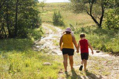 Yetişkin ve küçük bir çocuk, yemyeşil bir bitki örtüsü ve güneşli bir gökyüzü ile çevrili toprak bir yolda yürürken el ele tutuşuyorlar. Yolculukları hassas bir anı sergiliyor.