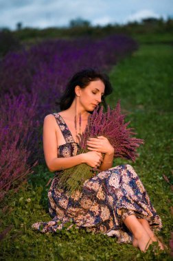 Canlı bir lavanta tarlasının ortasında elinde bir demet lavanta tutan bir kadının huzurlu görüntüsü. Altın saat ışığı sahneyi yıkıyor..