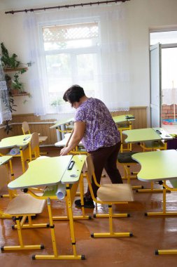 Bir kişi, güneşli bir sınıfta sarı okul sıralarını dikkatlice ayarlıyor. Masaların ve parlak ışığın düzenli bir şekilde düzenlenmesi ortamdaki öğrenme ve odaklanma için davetkar bir atmosfer yaratır..