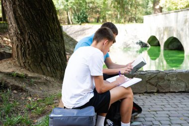 İki genç öğrenci, gölet ve köprüsü olan manzaralı bir parkta iş birliği yapıyorlar. Bir öğrenci bir kitaptan okurken diğeri not alıyor. Parkta ağaçlar var..