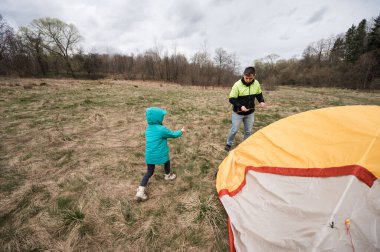 Bir baba ve kızı, yemyeşil ve vahşi doğada macera vaadiyle çevrili bulutlu bir gökyüzünün altında renkli bir çadır kurmak için birlikte çalışırlar..