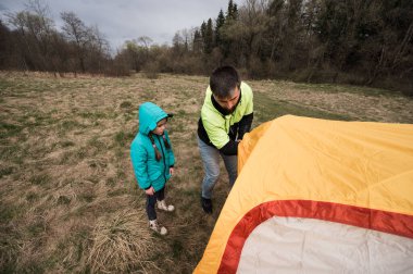 Bir adam ve bir kız açık otlak bir alanda canlı bir çadır kurmak için birlikte çalışırlar. Arkadaki ağaçlar dik duruyor, önlerindeki güzel bir kamp macerasına işaret ediyorlar..