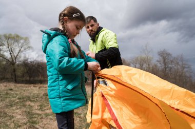 Bir baba, açık hava macerasına hazırlanırken kızına canlı bir çadır kurmasında yardım eder. Manzara bulutlu gökyüzüne ve tomurcuklanan ağaçlara sahiptir..
