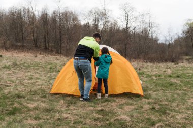 Neşeli bir baba, serin ve bulutlu bir günde ağaçlarla çevrili sakin bir çayırda parlak turuncu bir çadır kurarken kızına yardım eder..
