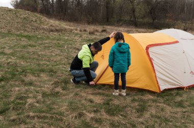 Turkuaz ceketli bir çocuk, bir yetişkinin yemyeşil çimenlerin üzerindeki sarı ve turuncu çadırı dikkatlice emniyete alıp birlikte açık hava maceralarını beklemesini izliyor..