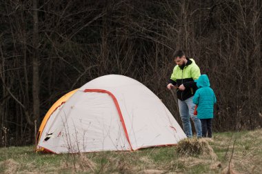İki birey, sakin bir ormanlık alanda rahat bir çadır kurarak akşam yaklaşırken doğanın harikaları arasında hoş bir kamp deneyimi için hazırlanıyorlar..