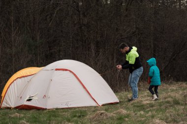 Bir baba ve çocuk, doğanın yeşillikleriyle çevrili, yumuşak, bulutlu gökyüzünün altında, huzurlu bir orman açıklığında, parlak bir çadır kurmak için birlikte çalışırlar..