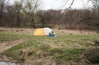 İki arkadaş, serin bir bahar gününde tomurcuklanan yeşillikler ve doğanın yumuşak sesleri ile çevrili huzurlu bir nehir kenarındaki canlı bir çadırın yanında dinleniyor..