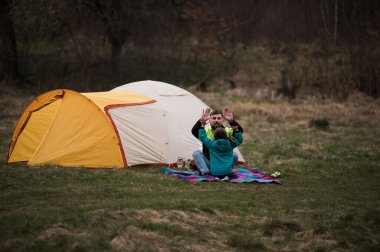 Bir çift çadırlarının önündeki renkli bir battaniyeye oturmuş, solgun ışıkta gülücükler ve şakacı hareketler yaparken, dışarıda neşeli bir toplantı gerçekleşiyor..