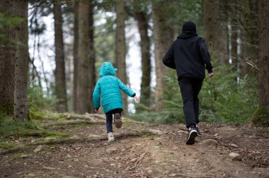 İki figür ormanlık bir patikadan geçiyor, parlak bir deniz mavisi ceket giyen bir çocuk yolu gösteriyor. Birlikte doğanın sessizliğinde neşeli anları paylaşıyorlar..