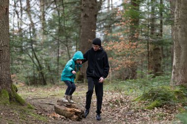 Huzurlu bir ormanda, bir çocuk güz renklerinin ılık ışıltısı ve yaprakların arasında şefkatli bir yetişkinin yardımıyla güvenli bir şekilde bir kütüğe basar..