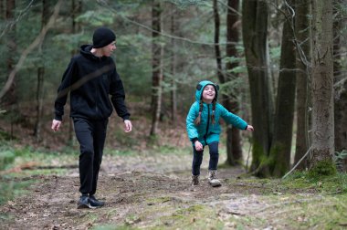 Parlak mavi ceket giymiş bir çocuk sevinçle öne doğru koşarken siyahlı bir yetişkin, uzun ağaçlar ve yumuşak orman zeminiyle çevrili canlı bir doğal ortamda onu yakından takip ediyor..