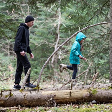 Canlı bir ormanın ortasında, bir çocuk düşmekte olan bir kütüğün üzerinde hevesle zıplarken, bir yetişkin arkasından yürüyor, doğayla bir anlık neşe ve bağ paylaşıyor..
