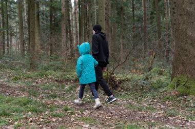 Parlak mavi paltolu bir çocuk, siyah kapüşonlu bir yetişkinin yanında yürür. Huzurlu bir orman patikasının içinden. Etrafı ağaçlarla çevrili ve yapraklarla kaplı..