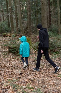 Bir ebeveyn ve çocuk, uzun ağaçlar ve düşmüş yapraklardan oluşan bir halı ile çevrili huzurlu bir orman yolunda geziniyor..