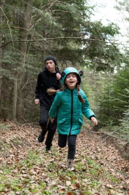 İki figür yapraklarla kaplı bir orman patikası boyunca neşeyle koşar, çocuk güler ve sıcacık giyinir, tabiat bahar zamanı canlı renklerle uyanır..