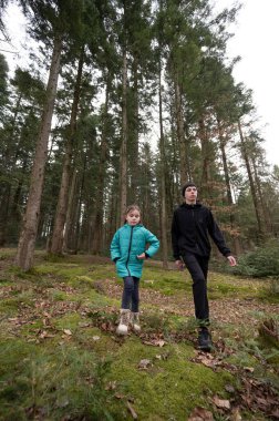 Parlak ceketli bir çocuk, uzun ağaçlar ve yosun halılarıyla çevrili ormanda bir yetişkinin yanında yürür ve doğadaki macera ruhunu yakalar..