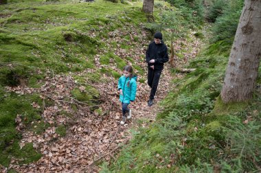 Parlak ceketli bir çocuk, yapraklarla kaplı sakin bir orman yolunda bir yetişkinin yanında yürür ve soğuk bir sonbahar gününde doğanın huzurunu kucaklar..