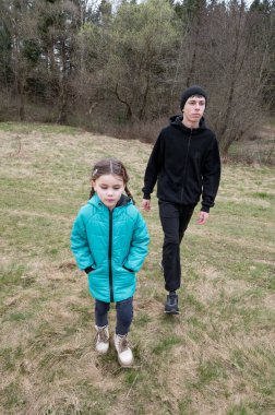 İki çocuk serin hava için sıcacık giyinmiş çimenli bir arazide birlikte yürüyorlar, sakin kırsal araziyi keşfederken eğleniyorlar..