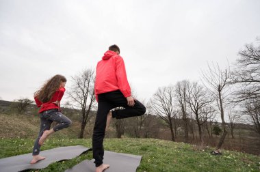 İki birey yoga minderleri üzerinde ısınma turuna çıkar. Ağaçlarla dolu yemyeşil, yemyeşil bir ortamda huzurlu bir sabahın tadını çıkarırlar..