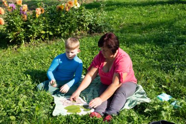 Güneşli bir günde dışarıda bir kadın ve genç bir çocuk çimenli bir tarlada oturup yaprakları ayırıyorlar. Battaniye önlerine serilmiş, yanında renkli çiçekler var..