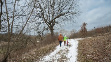 Bir çocuk, bulutlu gökyüzü ve yaprakları soyulmuş ağaçların altında sakin kış atmosferinin tadını çıkarırken, karlı bir patikada bisikleti itiyor..