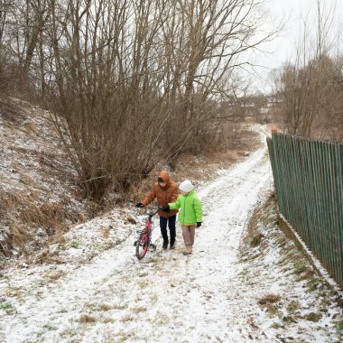 İki figür karla kaplı bir yolda yürüyor, biri serin bir kış gününde ağaçlarla çevrili bir halde bisikletli bir çocuğa rehberlik ediyor..