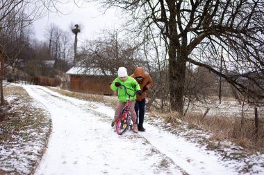 Bir çocuk karlı bir yolda bir yetişkinin yardımıyla bisiklet sürmeyi heyecanla öğrenir. Yakınlarda, rahat kulübeler ve yapraksız ağaçlar sakin bir kış ortamı yaratır..