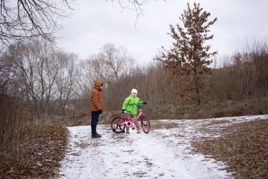İki çocuk, kar ve ağaçlarla dolu bir kış manzarasında oynarken, açık havada, biri parlak yeşil kıyafetli, diğeri de rahat kahverengi ceketli, eğlenceli bir an yaşıyor..