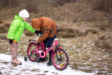 Youngsters engage in a fun winter activity as they work together to fix a vibrant pink bicycle, surrounded by soft snow and earthy tones of nature. clipart