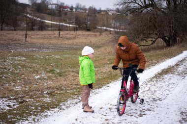 Biri parlak pembe bir bisiklet sürerken, diğeri yeşil giyinmiş, serin kış havasını heyecanlı bir şekilde izlerken, iki çocuk karlı bir tarlada şakalaşıyorlardı..