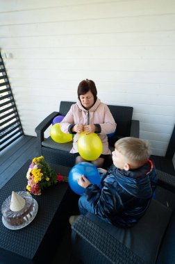 Bir kadın ve bir çocuk yaratıcı bir şekilde hazırlanırken parlak sarı ve mavi balonlar havayı dolduruyor. Çiçekler ve pastayla süslenmiş büyüleyici bir verandada neşeli bir kutlama..