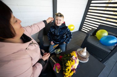 Bir kadın gülümseyen bir çocuğun kafasına bir pilot şapkası yerleştirirken keyifli bir doğum gününü renkli balonlarla süslenmiş rahat bir verandada kutlarken sevinç havayı doldurur..