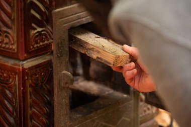 Dikkatlice bir odun parçasını eski bir sobaya yerleştirirken, eller, bir sıcaklık ve rahatlık geleneğiyle olan bağlantısını gösterir, kırsal bir ortamda sükunet anı somutlaştırır..