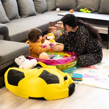 Parlak ve davetkar bir oturma odasında neşeli bir çocuk renkli oyuncakları keşfederken dikkatli bir yetişkin neşeli bir etkileşim içinde, sıcak bir bağ ve eğlence ortamı yaratıyor..