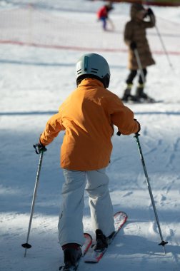 Parlak güneş, turuncu ceketli genç bir kayakçıyı karlı yamaçlarda dolaşırken aydınlatırken, diğer kayakçılar da tekniklerini uzaktan çalışırlar..