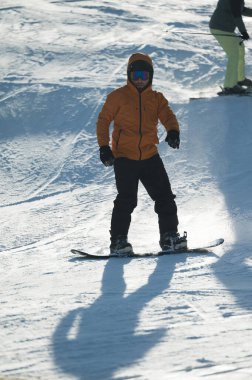 Parlak güneş ışığı, kar manzarasını aydınlatıyor. Turuncu giyinmiş yetenekli bir snowboardcu, yamaçtan aşağı doğru zahmetsizce süzülerek geride iz bırakıyor..
