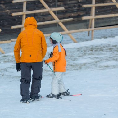 Açık mavi gökyüzünün altında bir ebeveyn, karlı kış harikalar diyarında ilk kayak deneyimlerine hazırlanırken küçük bir çocuğun kayak takımlarını giymesine yardım eder..
