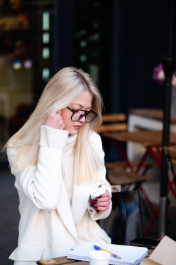 Düşünceli bir ifadeyle, şık giyinmiş şık bir kadın notlarını dengeler ve canlı bir kafe masasında bir tatlı, öğleden sonra güneşinin tadını çıkarır..