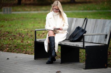 Şık bir ceket ve diz boyu çizmeler giyen genç bir kadın, sonbaharın canlı renkleriyle çevrili bir bankta güzel bir kitaptan hoşlanıyor..